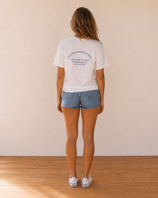 A woman viewed from behind wearing a white graphic t-shirt and denim shorts, standing on a wooden floor against a plain beige wall.