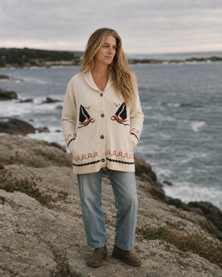 A woman stands on a rocky shoreline wearing a cream knit cardigan with nautical sailboat and wave motifs, light blue jeans, and brown boots, with the ocean and cloudy sky in the background.
