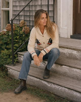 A woman sits on rustic wooden porch steps wearing a cream-colored cardigan with embroidered details, blue jeans, and dark boots in front of a house with gray siding.