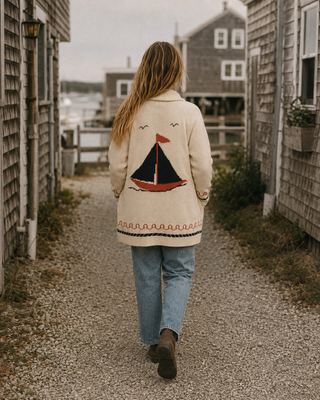 A woman viewed from behind walking down a gravel path between rustic wooden houses, wearing a cream-colored cardigan with a red and navy sailboat design on the back, paired with light blue jeans and brown boots.