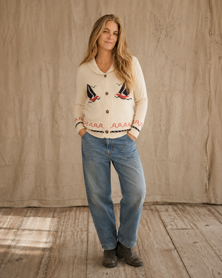 A woman stands in a rustic studio setting wearing a cream-colored button-up cardigan with embroidered sailboats and a braided trim, paired with blue jeans and dark boots.