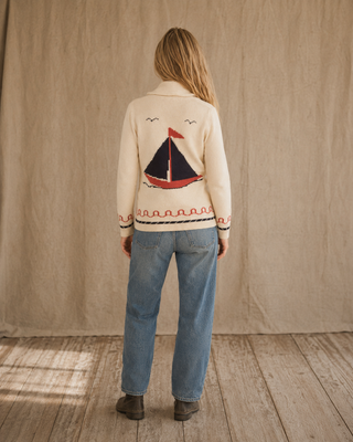 A woman viewed from behind wearing a cream-colored knit cardigan with a red and navy sailboat design on the back, paired with blue jeans and brown boots, standing against a neutral beige fabric backdrop.