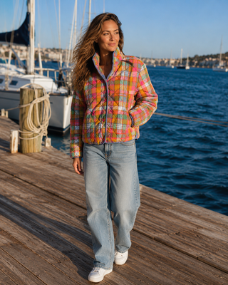 A woman stands on a wooden dock by the water wearing a colorful plaid zip-front jacket, light-wash jeans, and white sneakers. Boats and blue water are visible in the background.