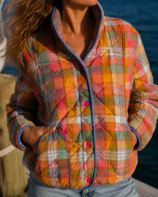 A close-up of a woman wearing a colorful quilted plaid jacket with a stand collar and pink buttons, set against a background of water and a dock.