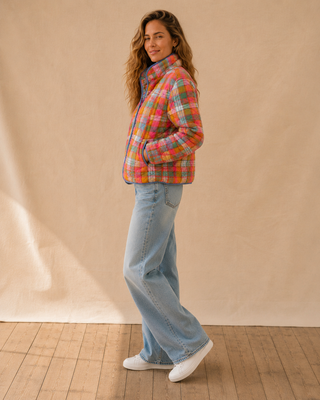 A woman stands in profile wearing a colorful plaid puffer jacket, light-wash wide-leg jeans, and white sneakers against a beige fabric backdrop and wood floor.