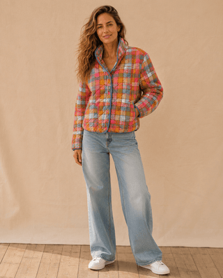 A woman modeling a colorful plaid zip-up jacket with a high collar, paired with light-wash wide-leg jeans and white sneakers, standing against a beige fabric backdrop.