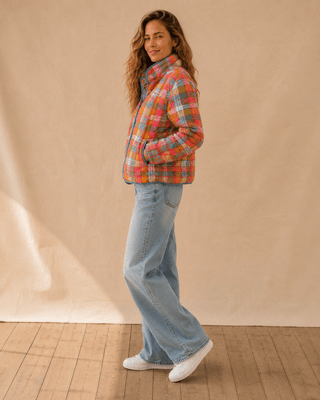 A woman stands in profile wearing a colorful plaid puffer jacket, light-wash wide-leg jeans, and white sneakers against a beige fabric backdrop and wood floor.