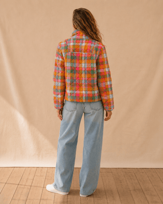 A person shown from the back wearing a colorful plaid button-up shirt and light blue wide-leg jeans, standing against a beige fabric backdrop on a wooden floor.