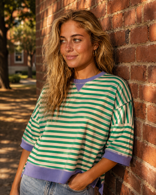 A woman with blonde hair leans against a red brick wall wearing a green and cream striped short-sleeve t-shirt with purple trim.