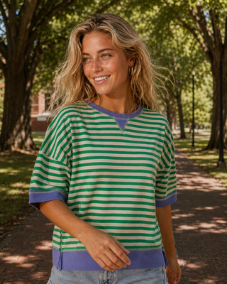 A woman wearing a green and white striped short-sleeve top with purple trim, standing on a tree-lined path outdoors.