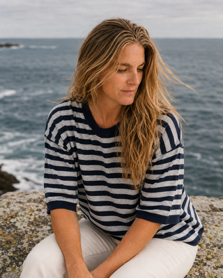 A woman sitting on a rocky cliff overlooking the ocean, wearing a navy and white horizontally striped short-sleeve top and white pants.