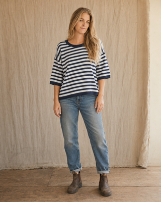 A woman stands against a textured beige fabric backdrop wearing a navy and white horizontally striped short-sleeve top, light-wash blue jeans, and dark brown ankle boots.