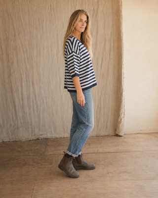 A woman stands in profile wearing a navy and white striped short-sleeve top, light blue jeans, and brown ankle boots against a neutral beige fabric backdrop.