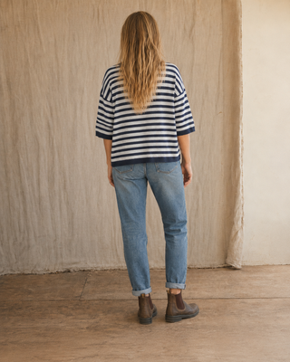 A woman viewed from behind wearing a navy and white striped short-sleeve top, light blue jeans, and brown Chelsea boots, standing against a textured beige wall and wooden floor.