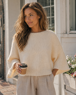 A woman wearing a cream-colored, textured knit poncho sweater and light beige pants, holding a takeaway coffee cup in front of a white building with windows.