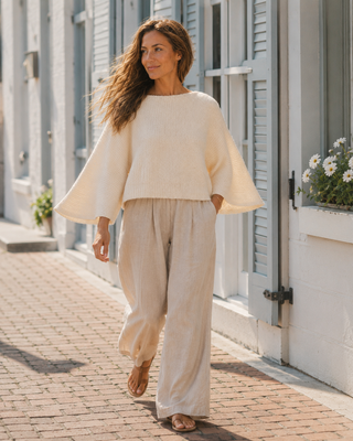 A woman walking outdoors on a brick sidewalk beside white buildings with gray shutters, wearing a cream ribbed knit long-sleeve top with wide bell sleeves and matching cream wide-leg pants.