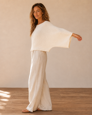 A woman modeling a cream-colored ribbed knit poncho-style top and matching wide-leg pants in a minimalist studio setting with a beige wall and wooden floor.