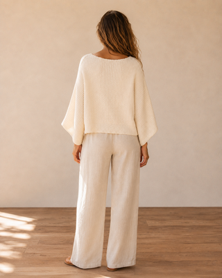A woman viewed from behind wearing a cream-colored ribbed knit top with wide draped sleeves and matching cream wide-leg pants, standing on a wooden floor against a neutral beige wall.