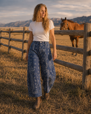 A woman stands in a sunlit grassy field beside a wooden fence, wearing a white short-sleeve top and loose blue patterned wide-leg pants, with a horse and mountains in the background.