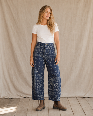 A woman stands in front of a beige fabric backdrop wearing loose dark blue paisley pants, a white short-sleeve T-shirt, and brown ankle boots.