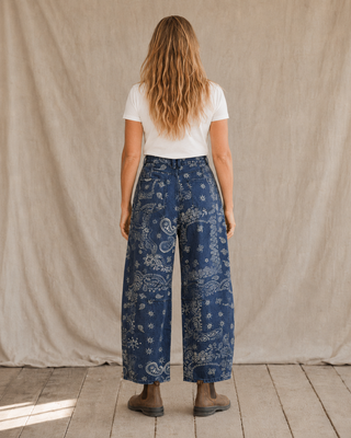 A woman stands with her back to the camera wearing blue wide-leg pants with a white paisley and floral bandana print, a white t-shirt, and brown boots, set against a neutral beige fabric backdrop.