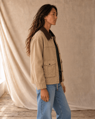 A woman wearing a beige canvas utility jacket with a dark brown corduroy collar, styled with light blue jeans and a black top, standing in front of a draped beige fabric backdrop.