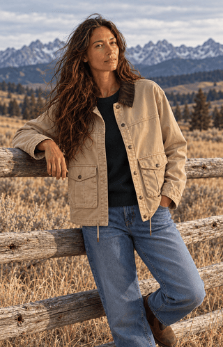 A woman wearing a beige utility jacket with a dark brown corduroy collar, blue jeans, and a black top, leaning against a rustic wooden fence in a mountain landscape.