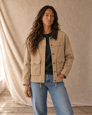 A woman modeling a beige utility jacket with a dark brown corduroy collar, worn over a dark green t-shirt and blue jeans, standing against a draped beige fabric backdrop.