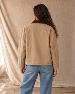 A woman shown from the back wearing a beige utility jacket with a dark brown collar and light blue jeans, standing in front of a draped beige fabric backdrop.