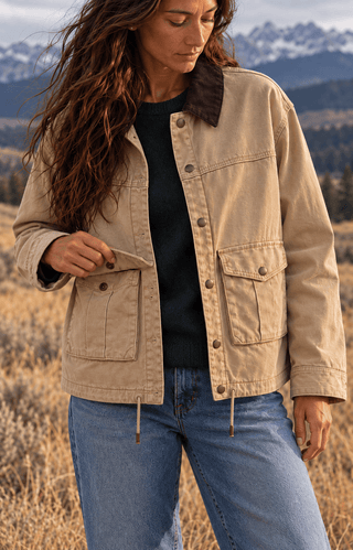 A woman wearing a beige canvas utility jacket with a dark brown corduroy collar, layered over a black top and blue jeans, standing in a dry grassy field with mountains in the background.