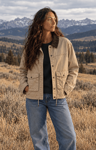 A woman stands outdoors in a dry grassy field with mountains in the background, wearing a beige button-front jacket with a dark brown collar, a black top, and blue jeans.